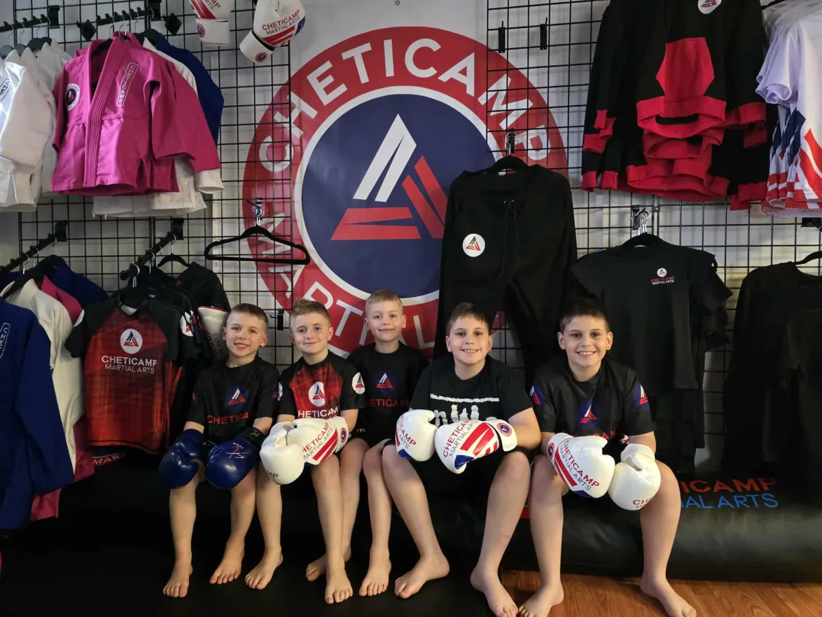 Children sit barefoot in martial arts gear beneath a red-and-black academy logo, with uniforms displayed behind them.