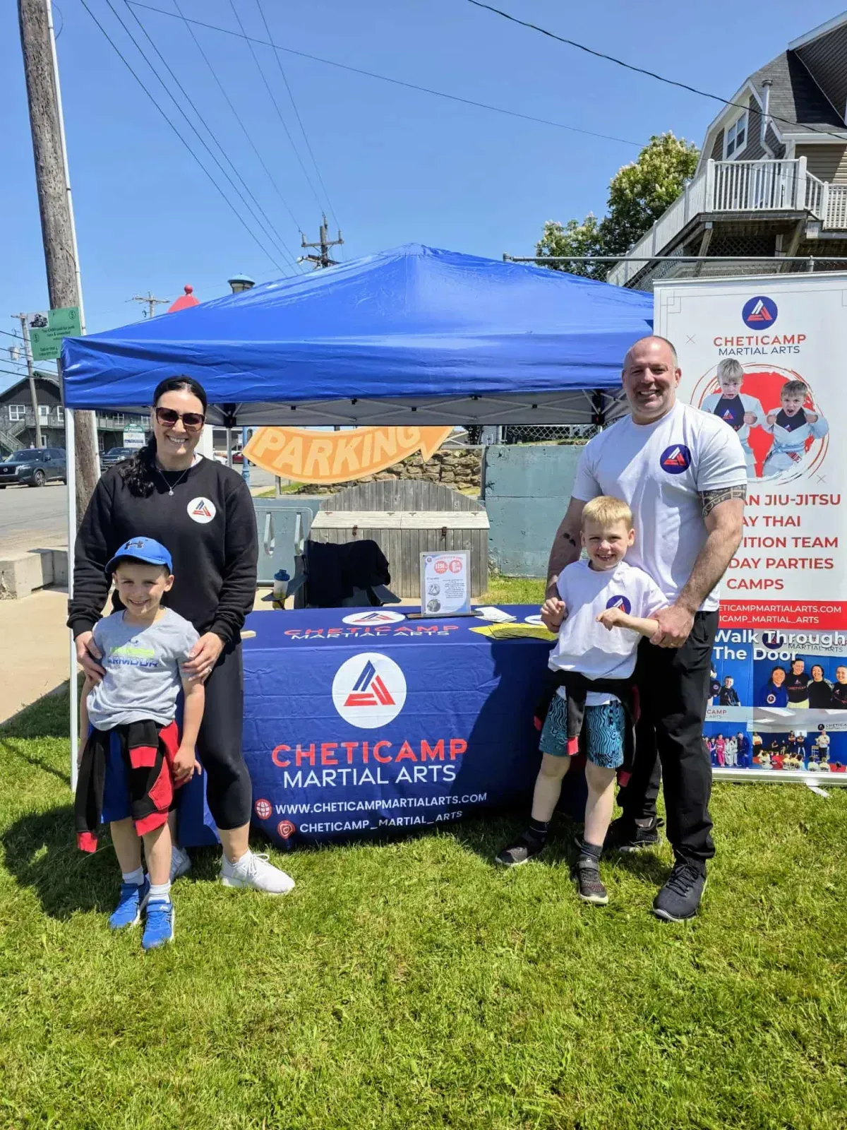Three people standing by a blue martial arts camp booth at an outdoor event, holding certificates.