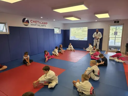 Children sitting on mats in a martial arts studio, with an instructor standing at the front.
