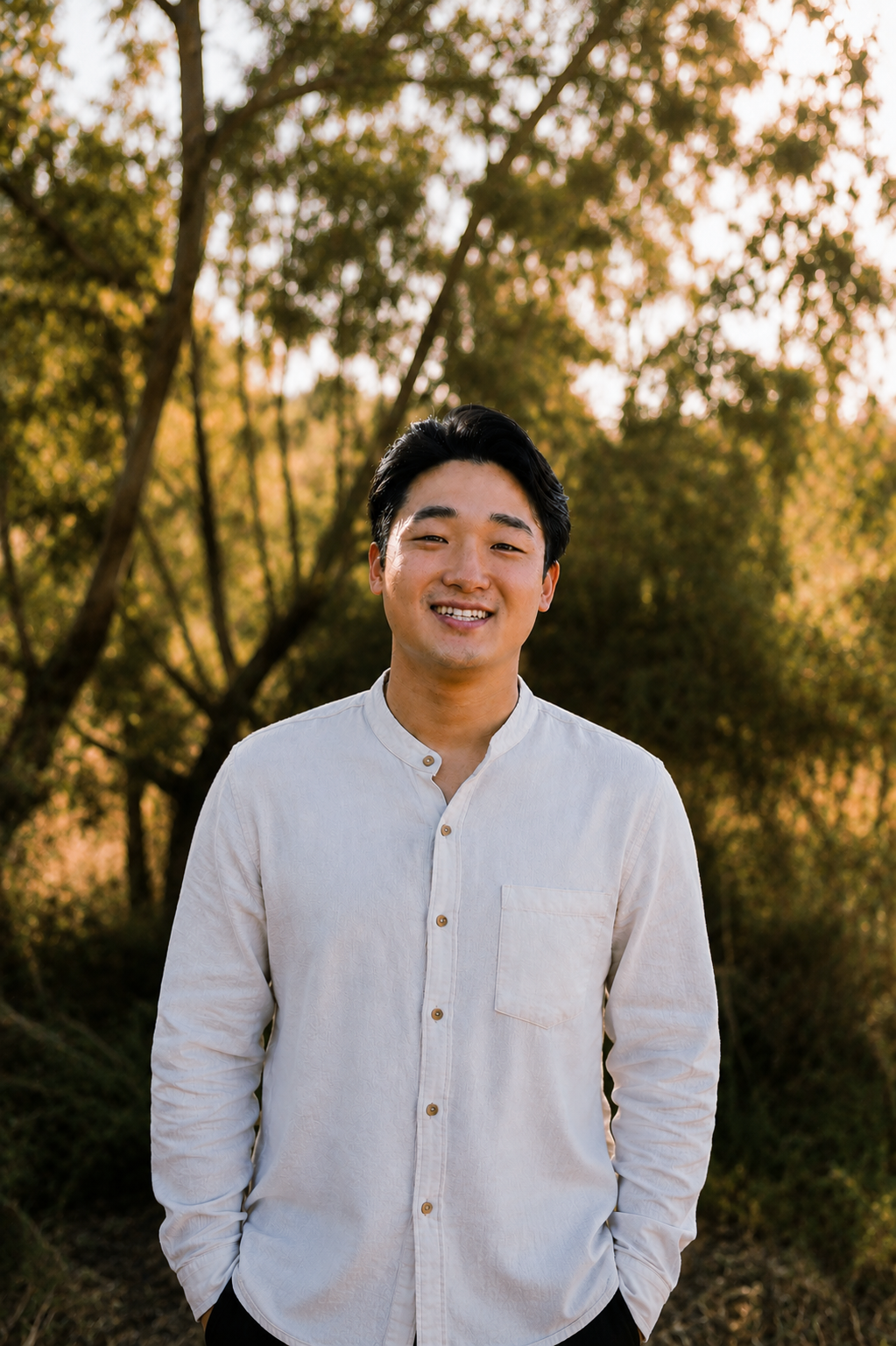 Smiling person in a white button-up shirt standing outdoors among sunlit trees.