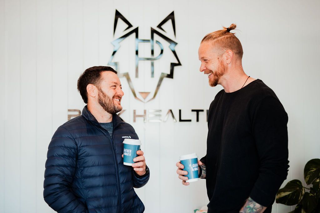 Two people smiling and chatting while holding cups in front of a wall logo