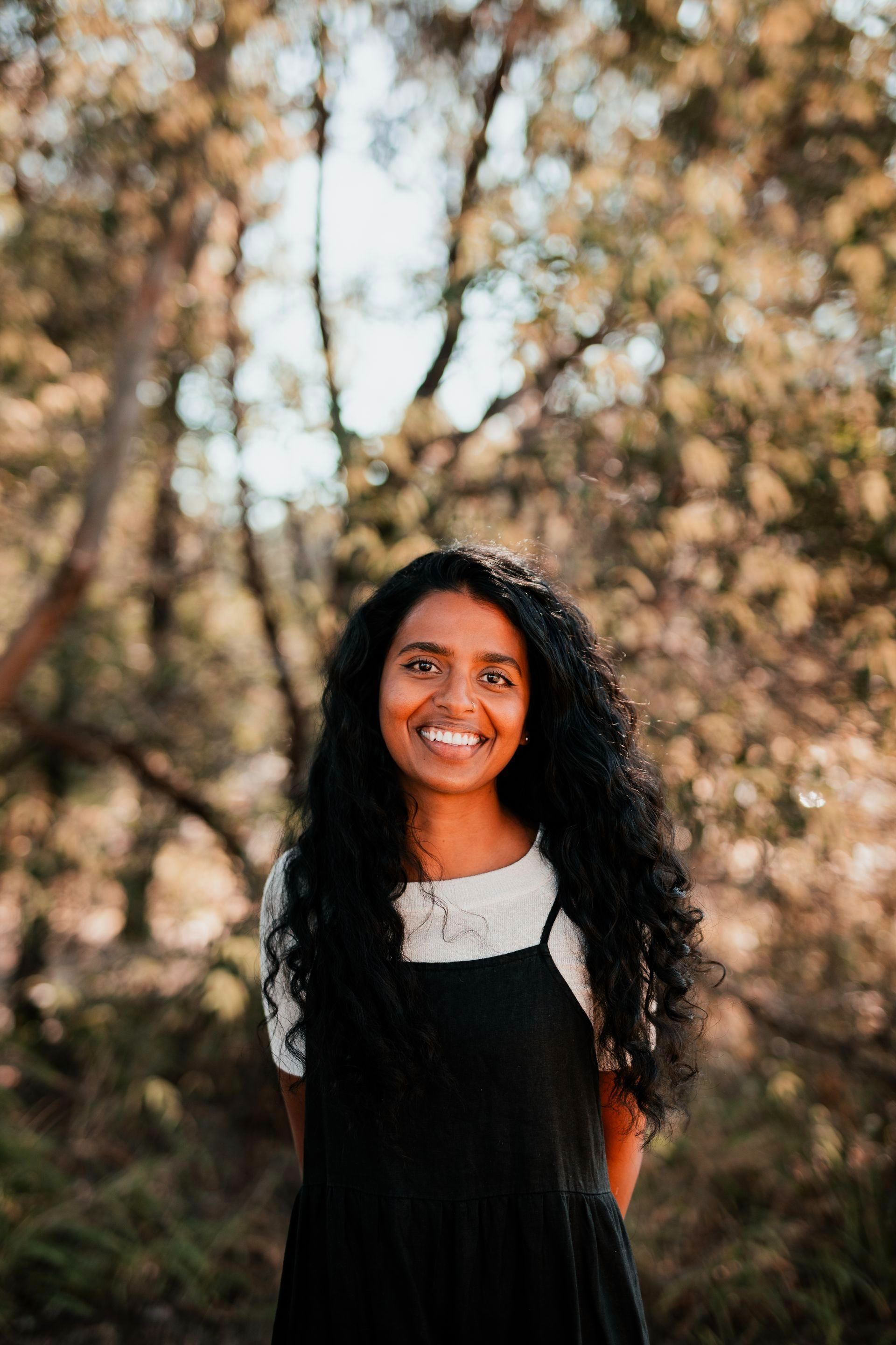 Smiling person in a black dress standing outdoors among trees with a blurred background