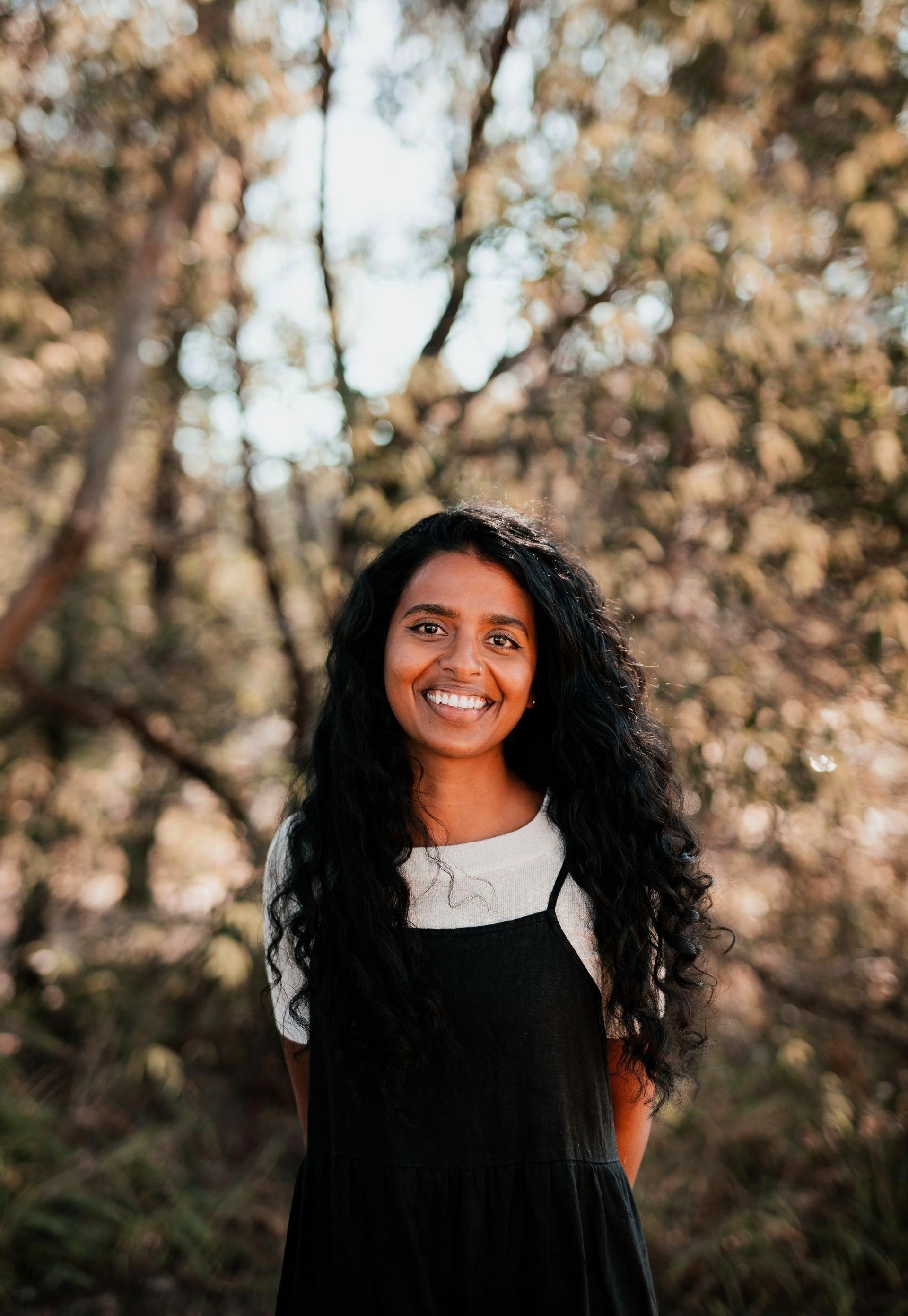 Woman smiling outdoors in a black dress and white top, standing among trees.