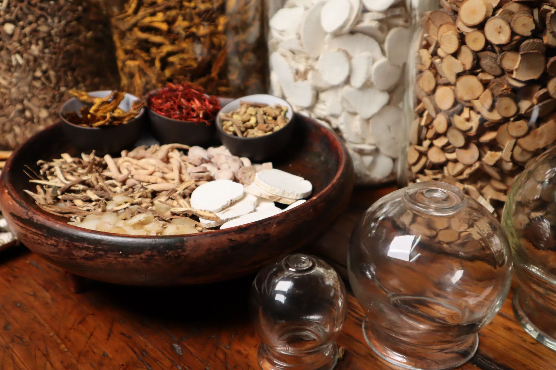 A bowl of dried herbs and roots beside glass jars and mushrooms on a wooden table