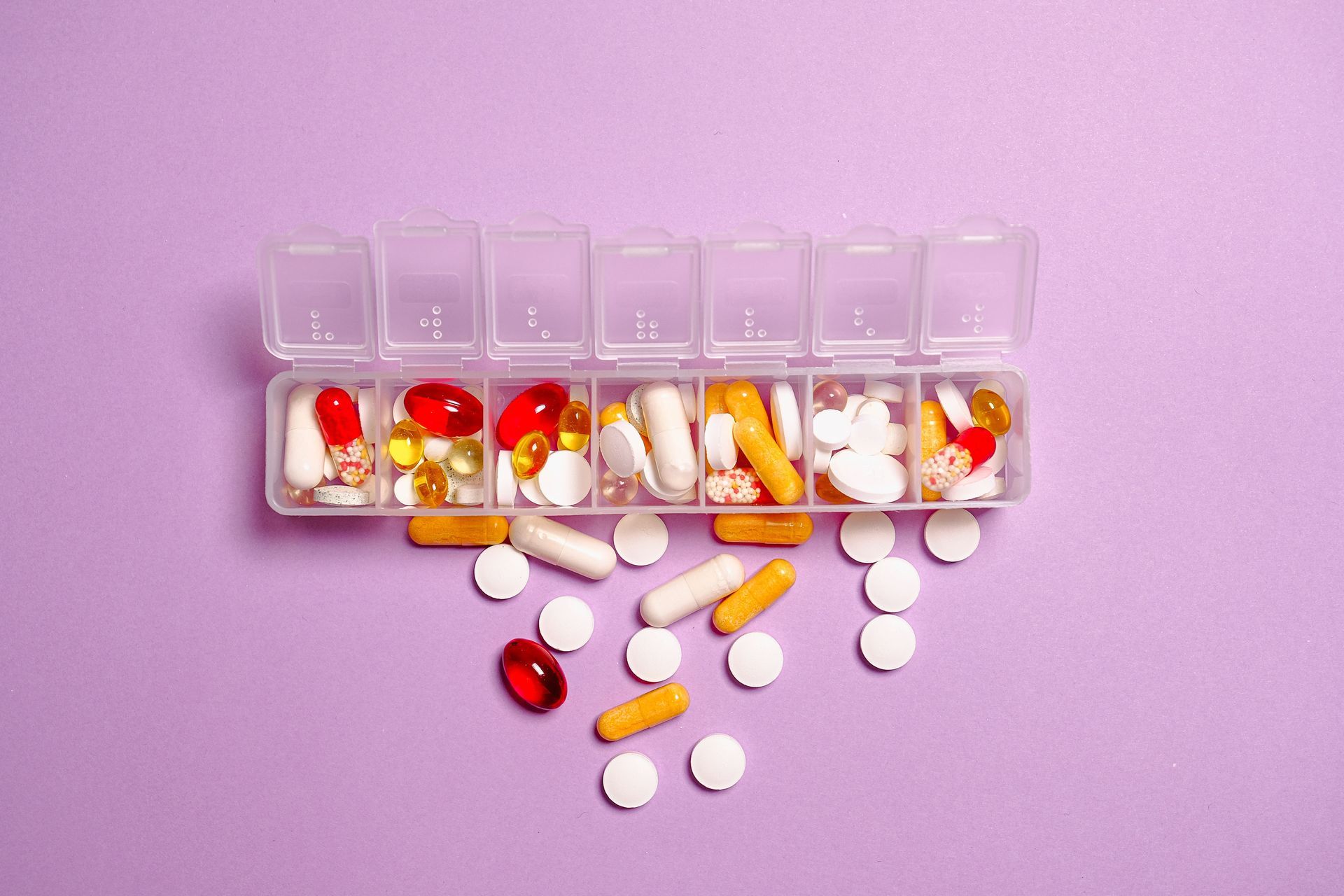 Weekly pill organizer with assorted colorful pills spilled onto a purple surface