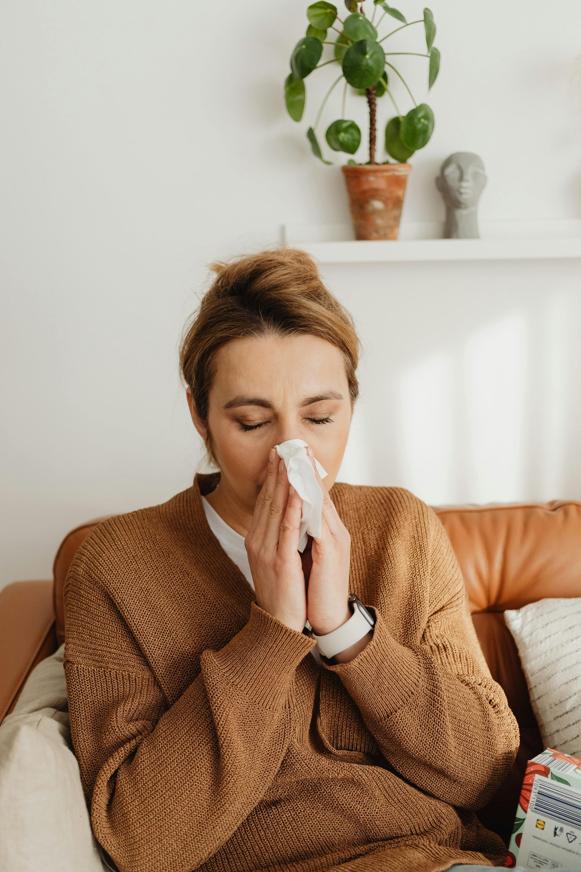 Woman on couch blowing her nose with a tissue in a cozy living room