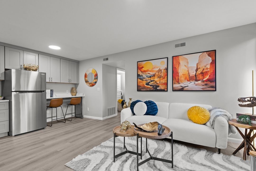 Modern living room with white sofa, coffee table, wall art, and small kitchen area with stainless steel refrigerator