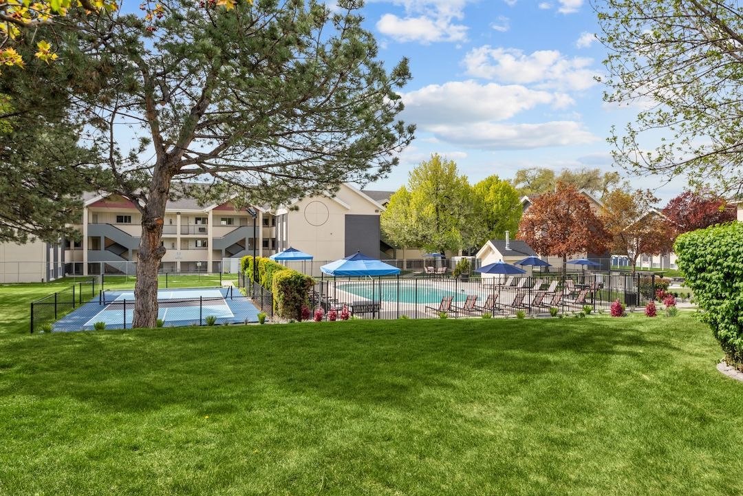 Apartment complex pool area with blue umbrellas, trees, and green lawn under a partly cloudy sky