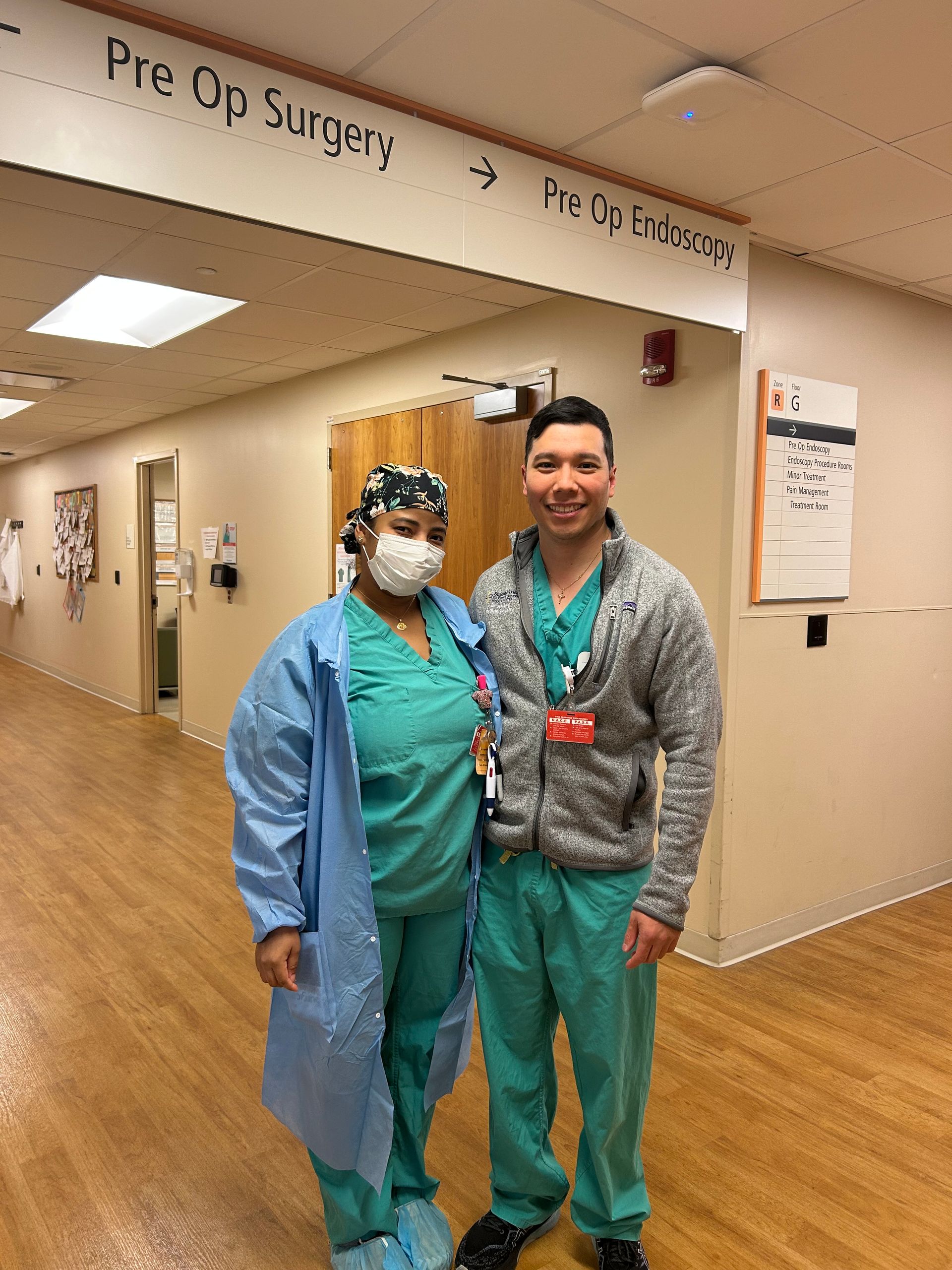 Two hospital staff in scrubs pose in a pre-op surgery hallway, one wearing a mask.