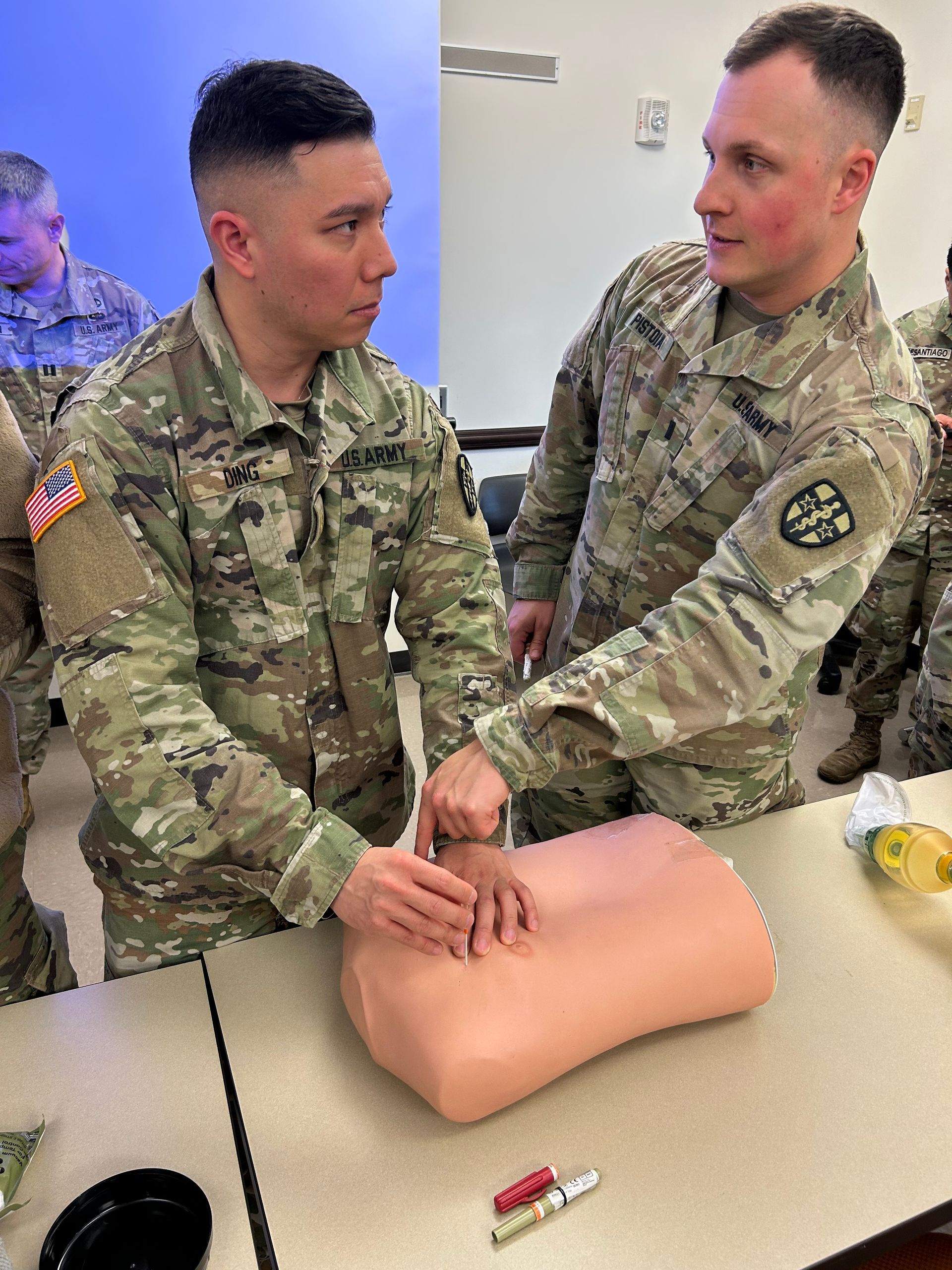 Two soldiers practice CPR on a training dummy indoors. One soldier guides the other's hands.