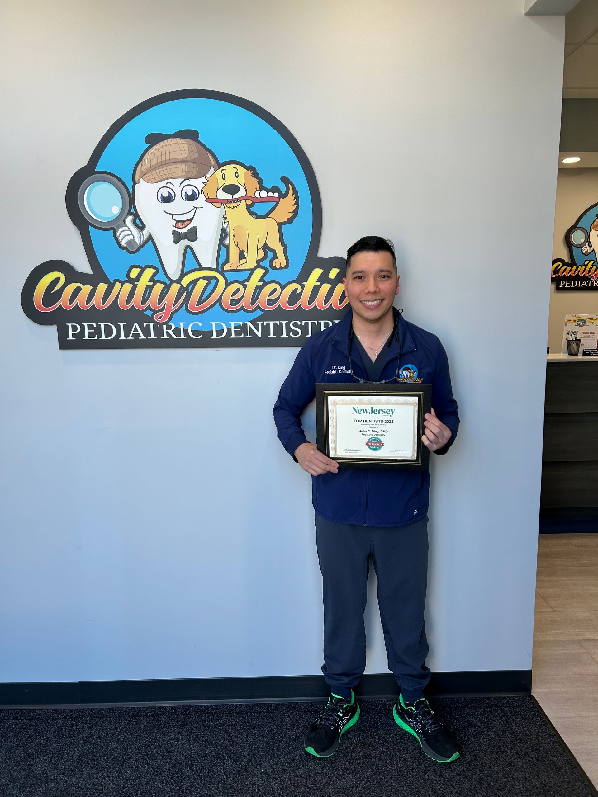 Man holding an award stands in front of a pediatric dentist office logo. The logo features a tooth, magnifying glass and dog.
