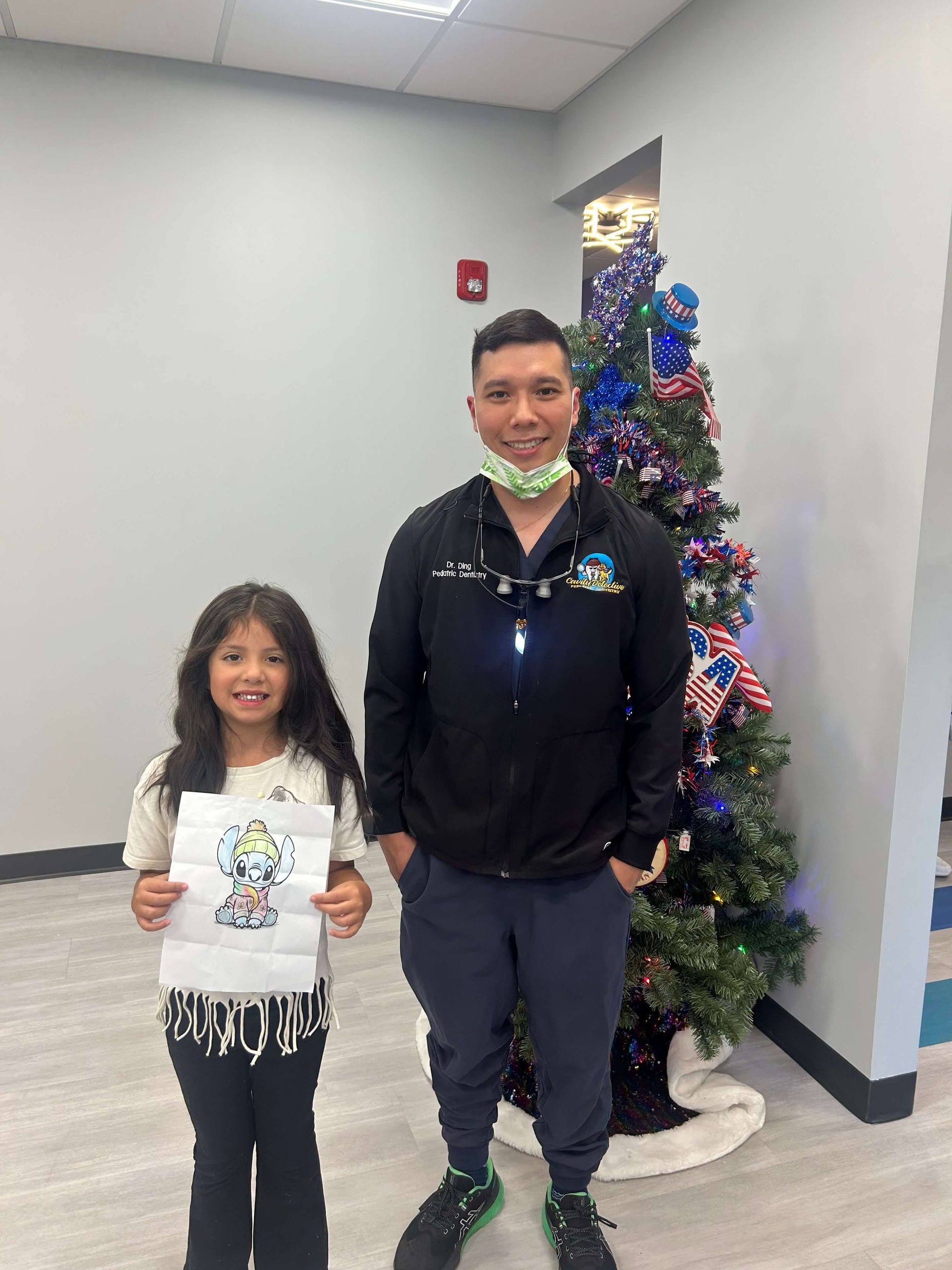 Two people stand smiling indoors beside a decorated Christmas tree.