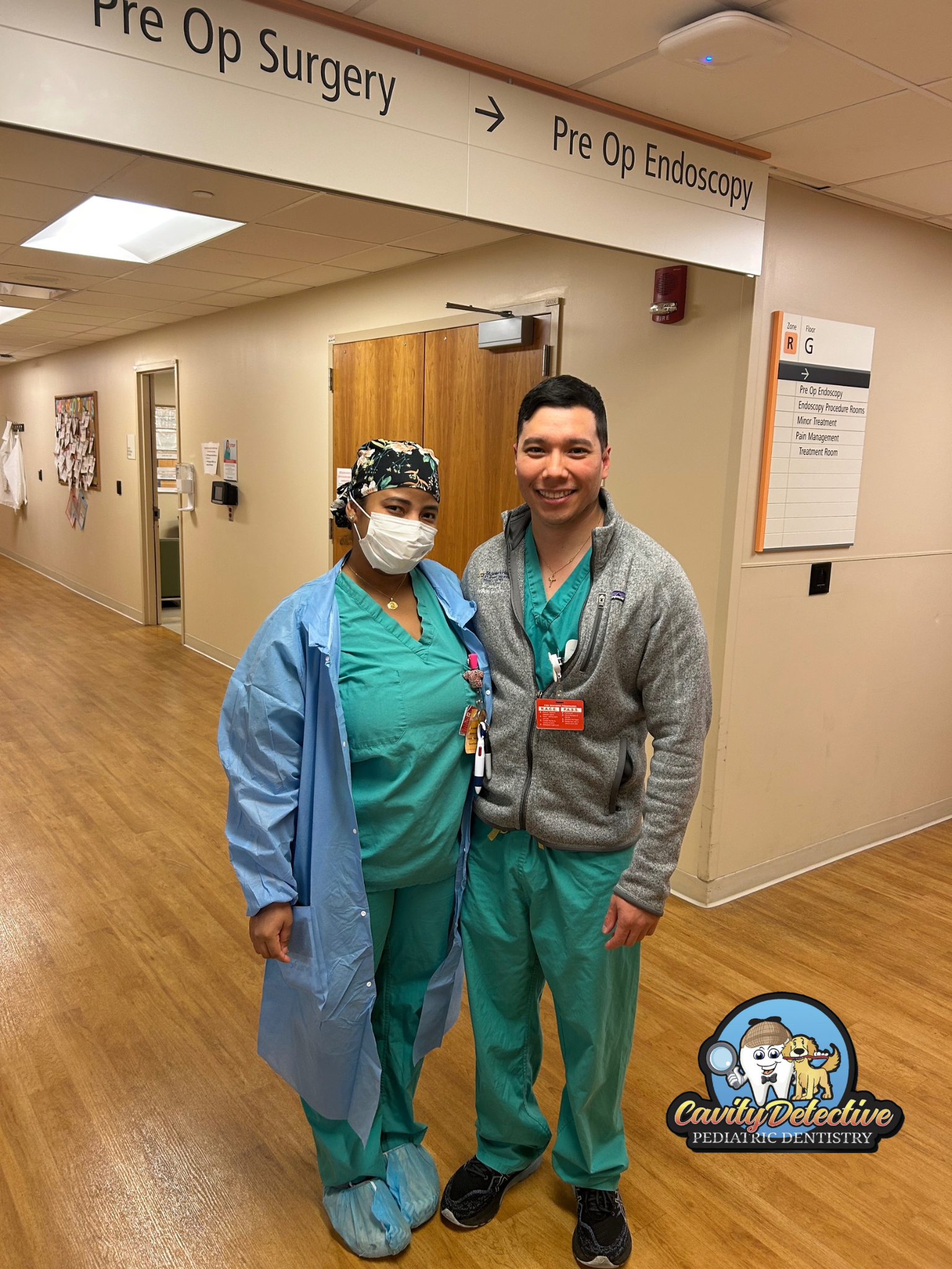 Two medical professionals in scrubs pose in hospital hallway; a sign points to Pre-Op Surgery.