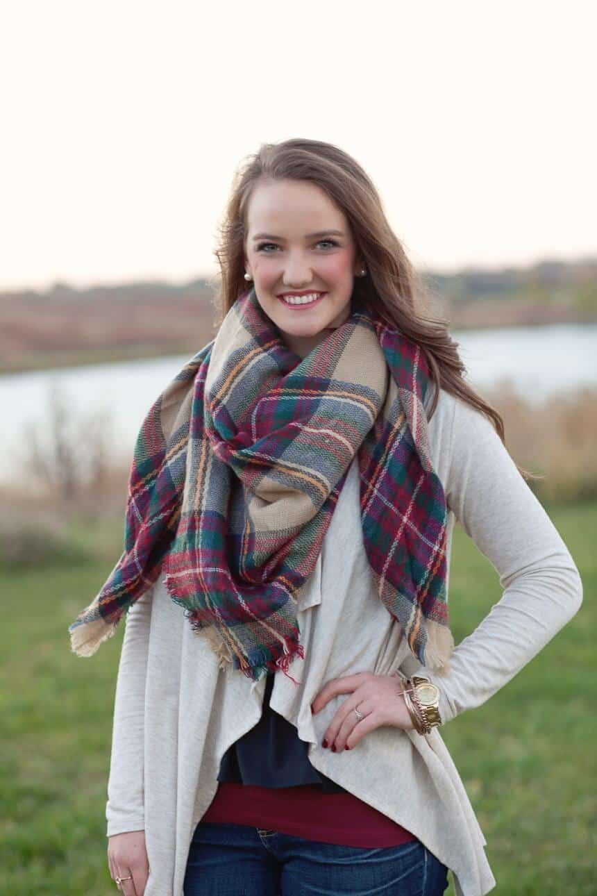 A woman wearing a plaid scarf and a white cardigan is standing in a field.