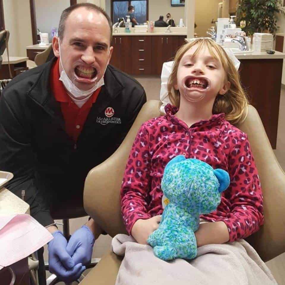 A little girl is sitting in a dental chair holding a teddy bear