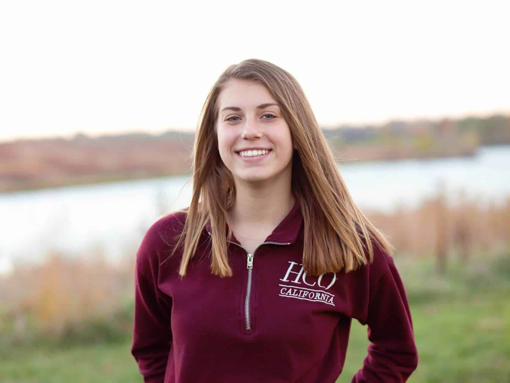 A young woman wearing a maroon sweatshirt is standing in front of a body of water.