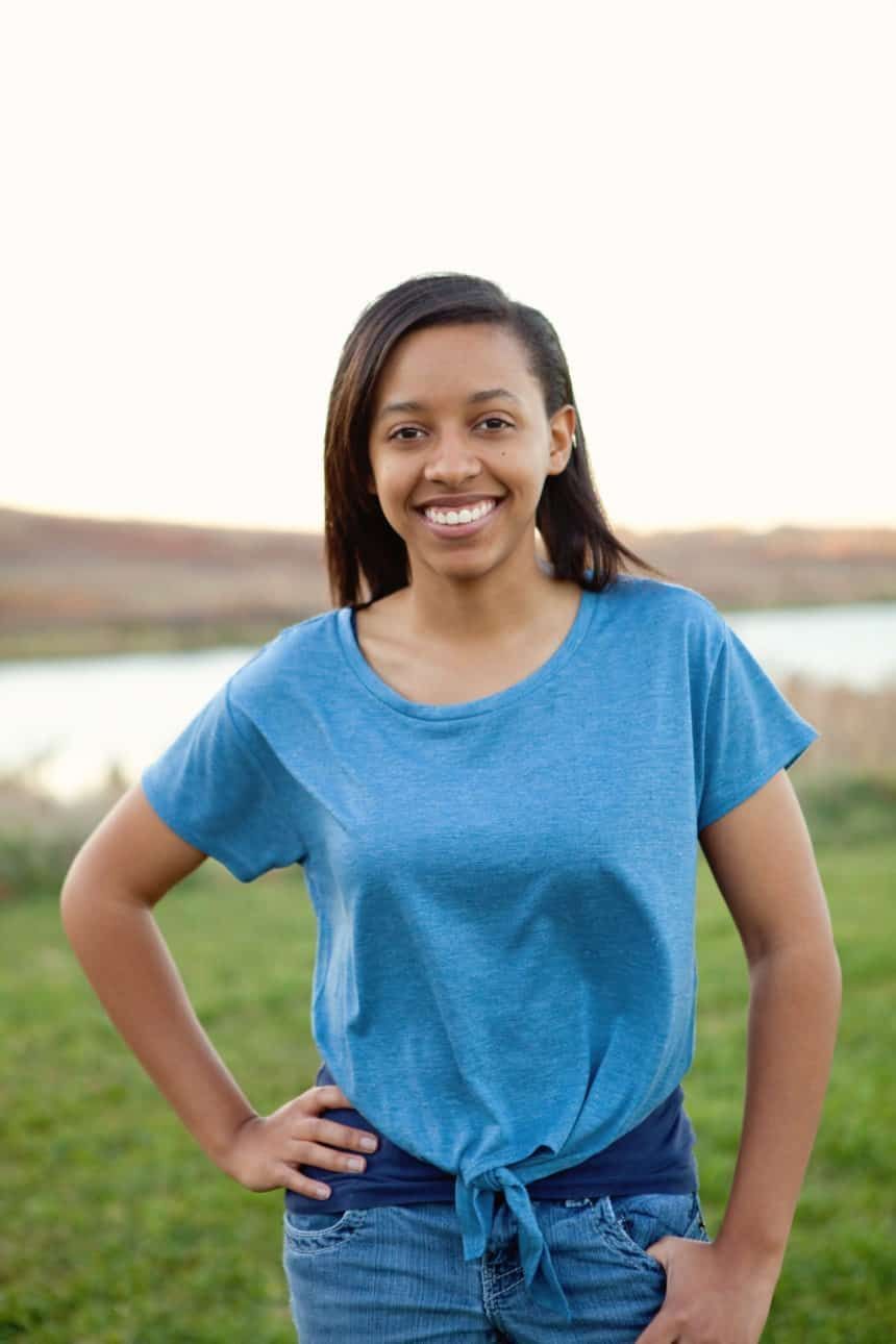 A woman in a blue shirt and jeans is standing in a field with her hands on her hips.