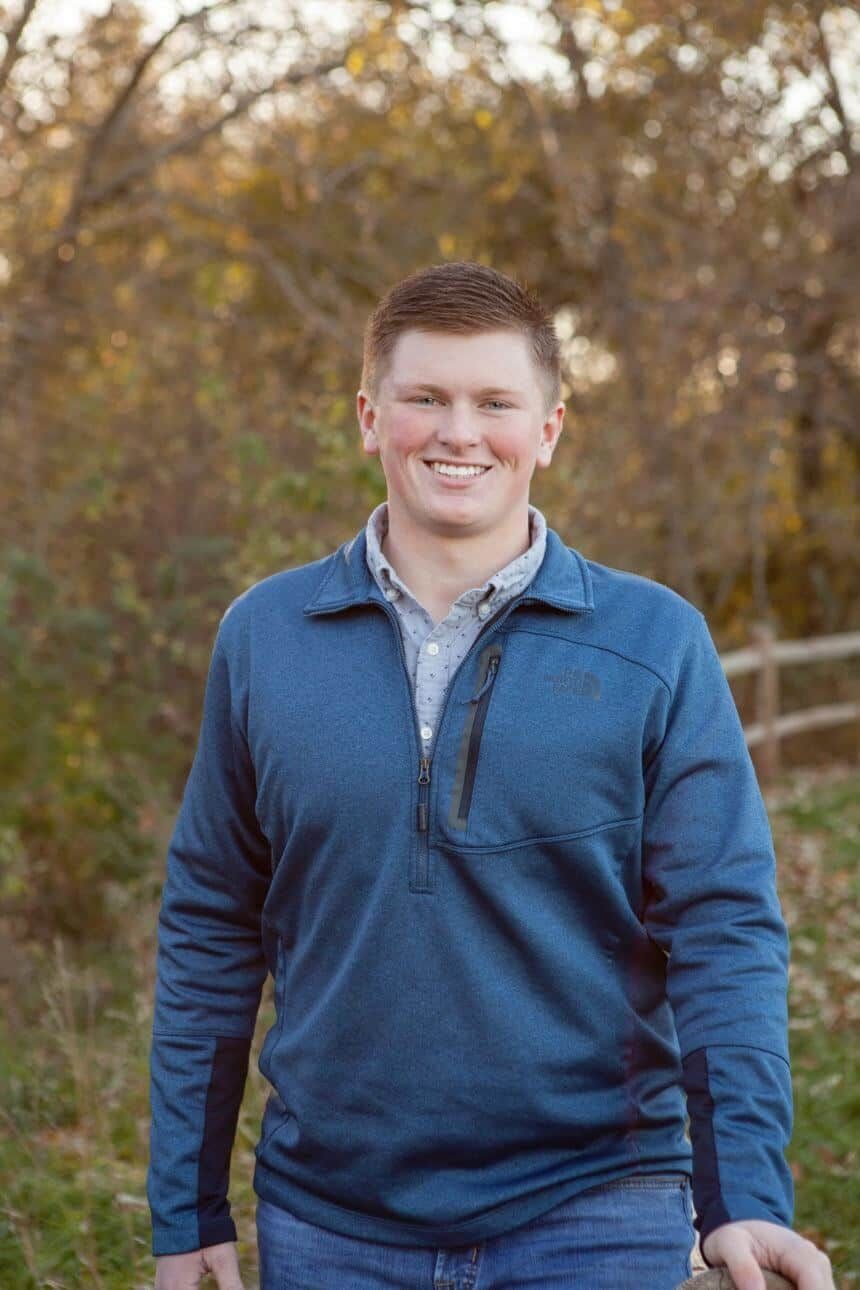 A man in a blue sweater is standing in front of a fence.
