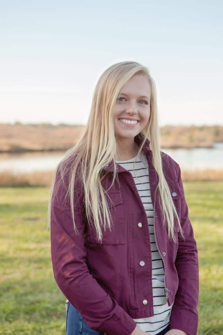 A young woman wearing a purple jacket and a striped shirt is standing in a field.