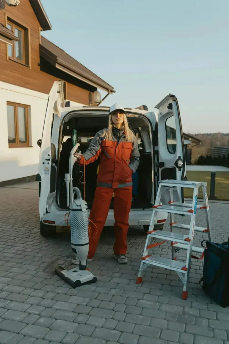 Person in orange coveralls stands by open van doors beside a step ladder and vacuum on a driveway.
