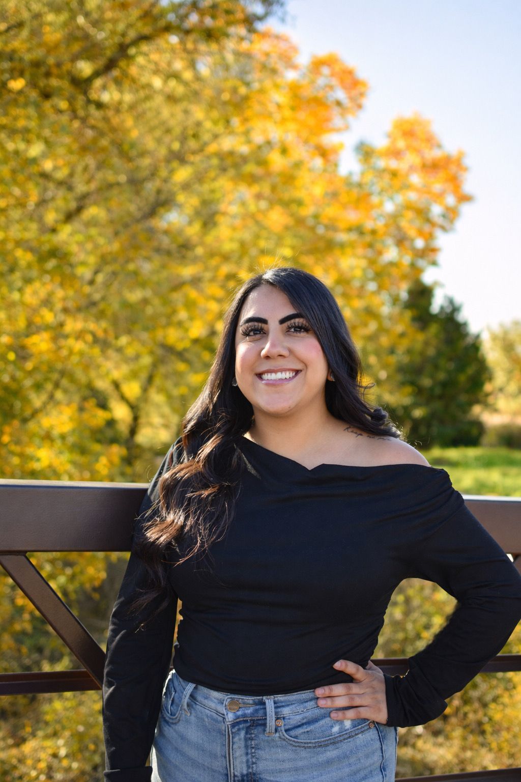 Smiling woman in a black top and jeans standing on a wooden bridge with autumn trees behind her