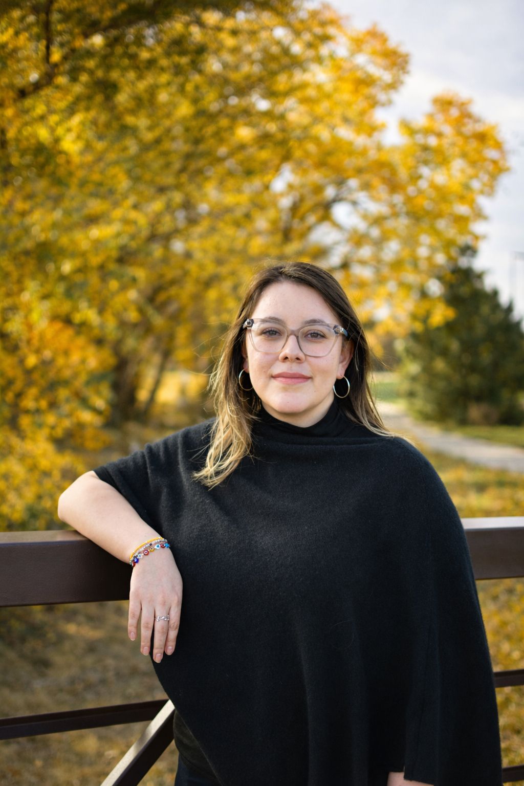 Person leaning on a wooden railing outdoors in a black top, with autumn trees in the background