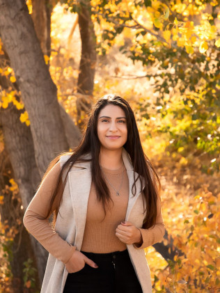 Woman in a tan sweater and vest standing outdoors among autumn trees, smiling at the camera.