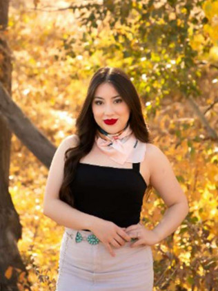 Woman in a black dress and pale pink scarf stands outdoors among autumn leaves.