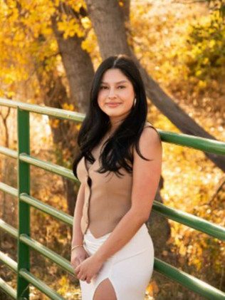 Woman standing by a green railing in autumn, wearing a beige top and white pants, with yellow foliage behind her
