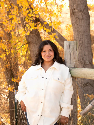 Smiling person in a white jacket standing among trees with golden autumn leaves