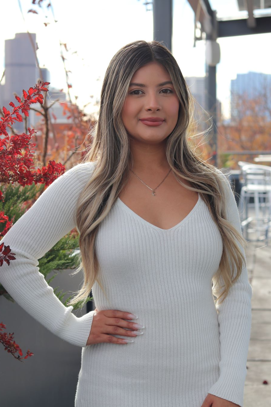 Woman in a white sweater standing outdoors by red flowers, smiling with one hand on her hip.