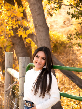 Woman in a white sweater smiling by a green fence in a sunlit autumn park.
