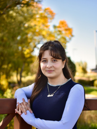 Young woman standing outdoors with arms crossed, wearing a navy dress over a white shirt, with autumn trees behind her