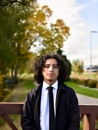 Person in a black jacket and tie standing outdoors in a park with autumn trees.