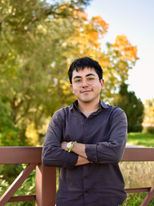 Person standing with arms crossed on a wooden bridge, wearing glasses and a dark shirt, with autumn trees behind.