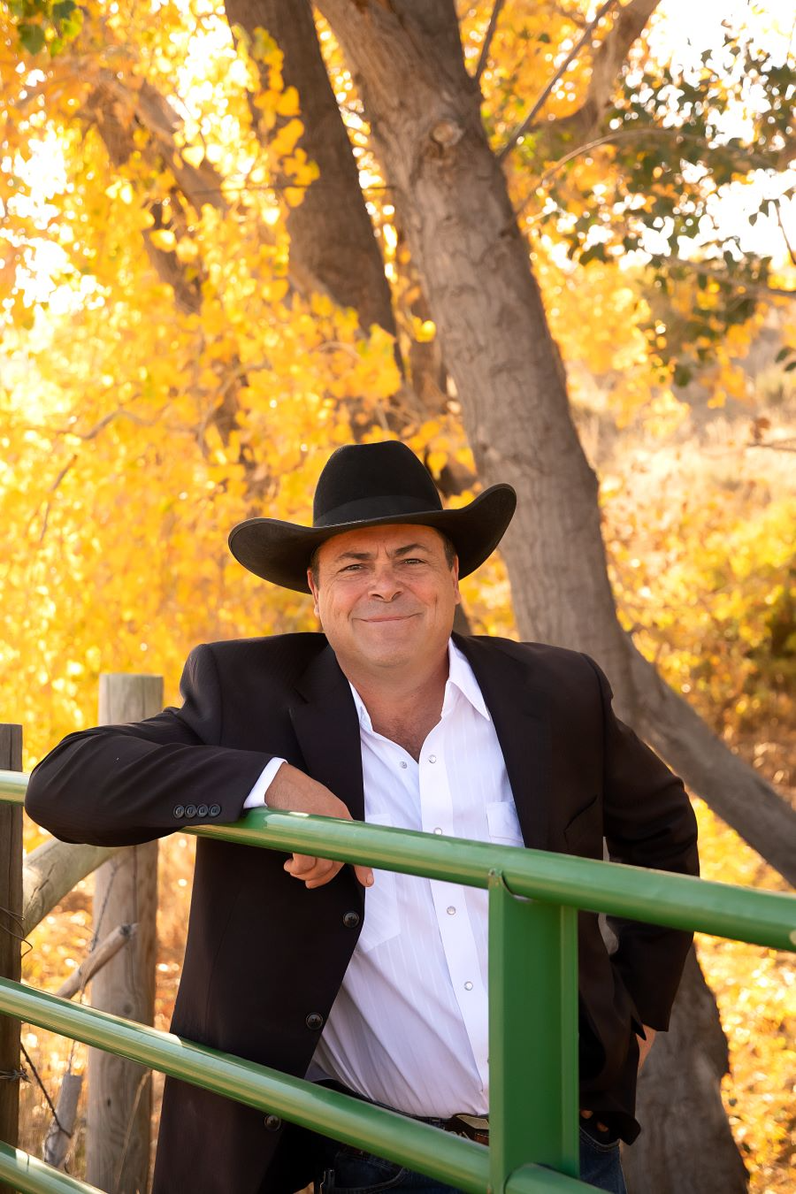 Man in black cowboy hat leaning on green railing amid golden autumn trees