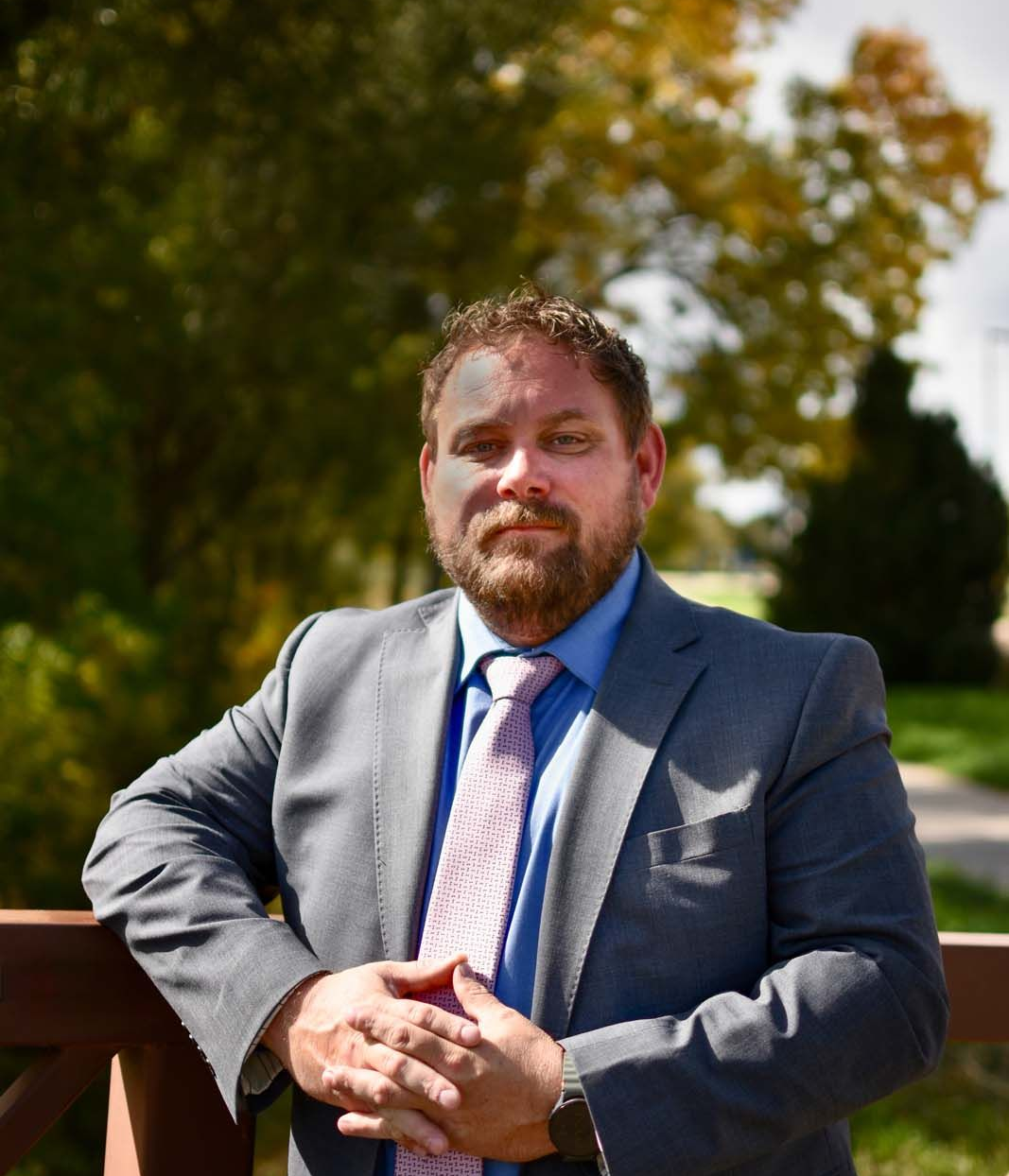 Man in a gray suit and blue shirt standing outdoors by a wooden railing in sunlight