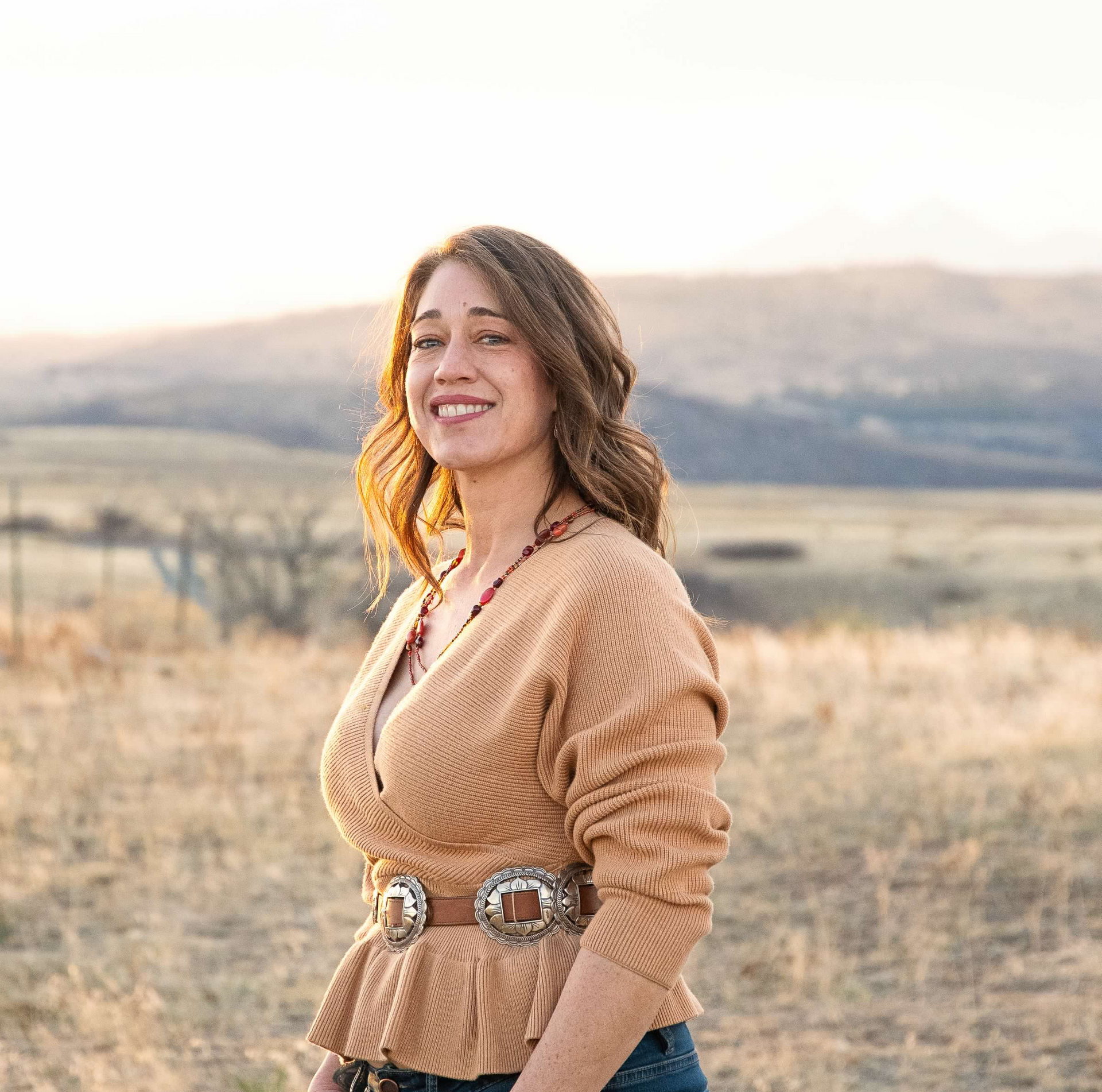 Smiling woman in a tan sweater standing in a sunlit grassy field with hills in the background