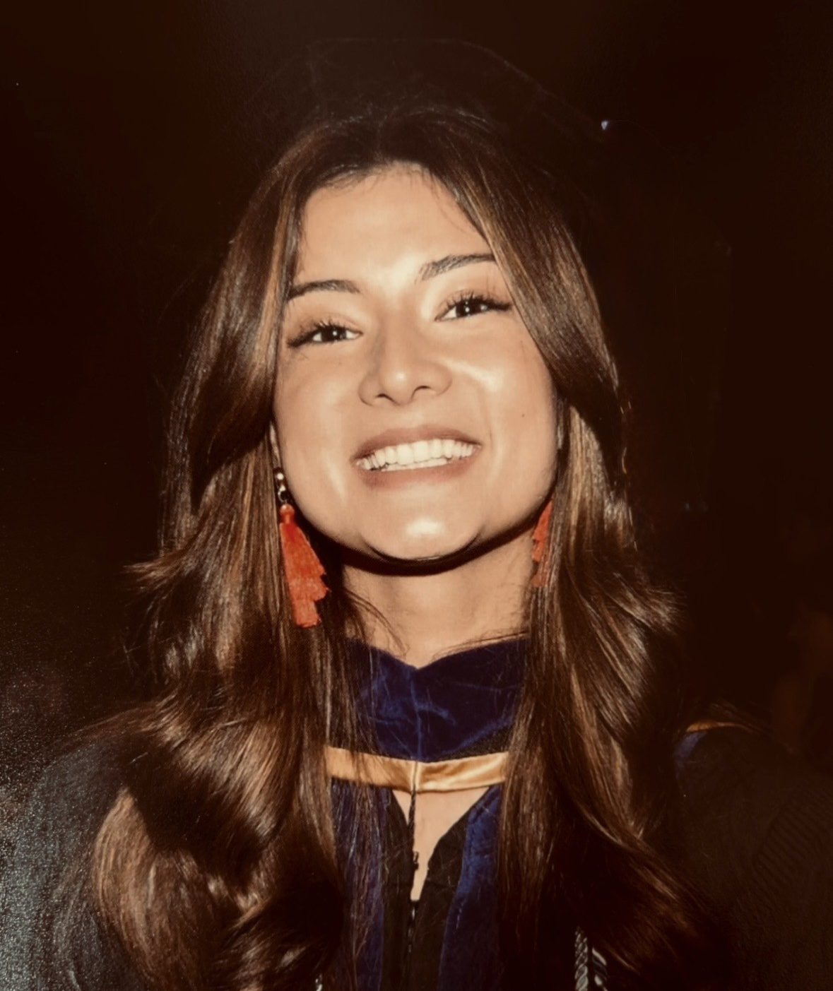Smiling woman with long wavy hair, red earrings, and a dark outfit against a black background