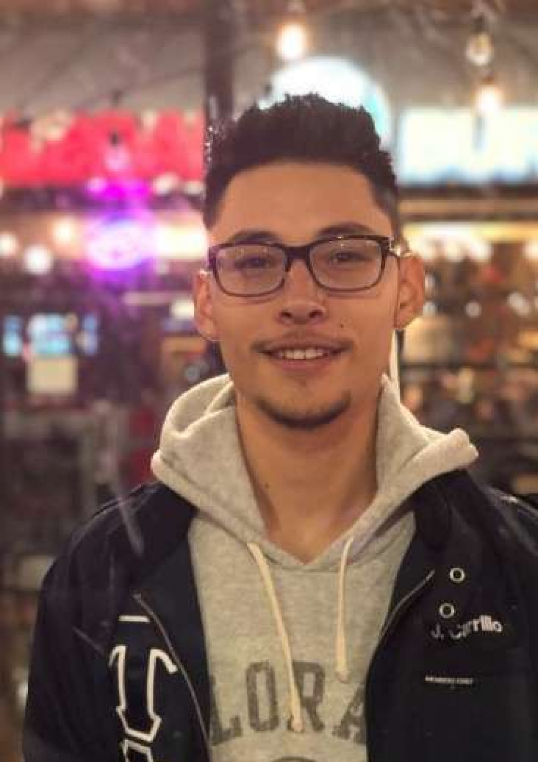 Smiling person wearing glasses and a hoodie, standing in a softly lit indoor setting with colorful lights.