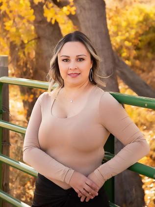 Woman leaning on a green railing outdoors among autumn trees, wearing a beige top and smiling.