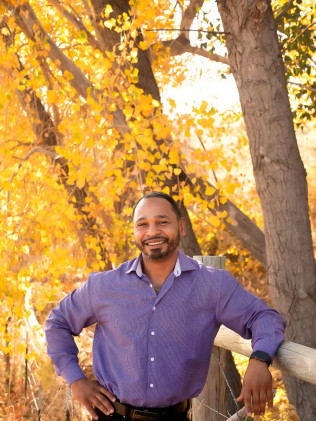 Smiling man in a purple shirt standing by a tree with bright yellow autumn leaves