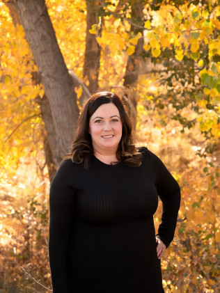 Woman in a black dress standing outdoors among golden autumn trees, smiling at the camera.