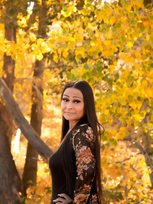Woman in a black dress standing outdoors among golden autumn leaves, looking at the camera.