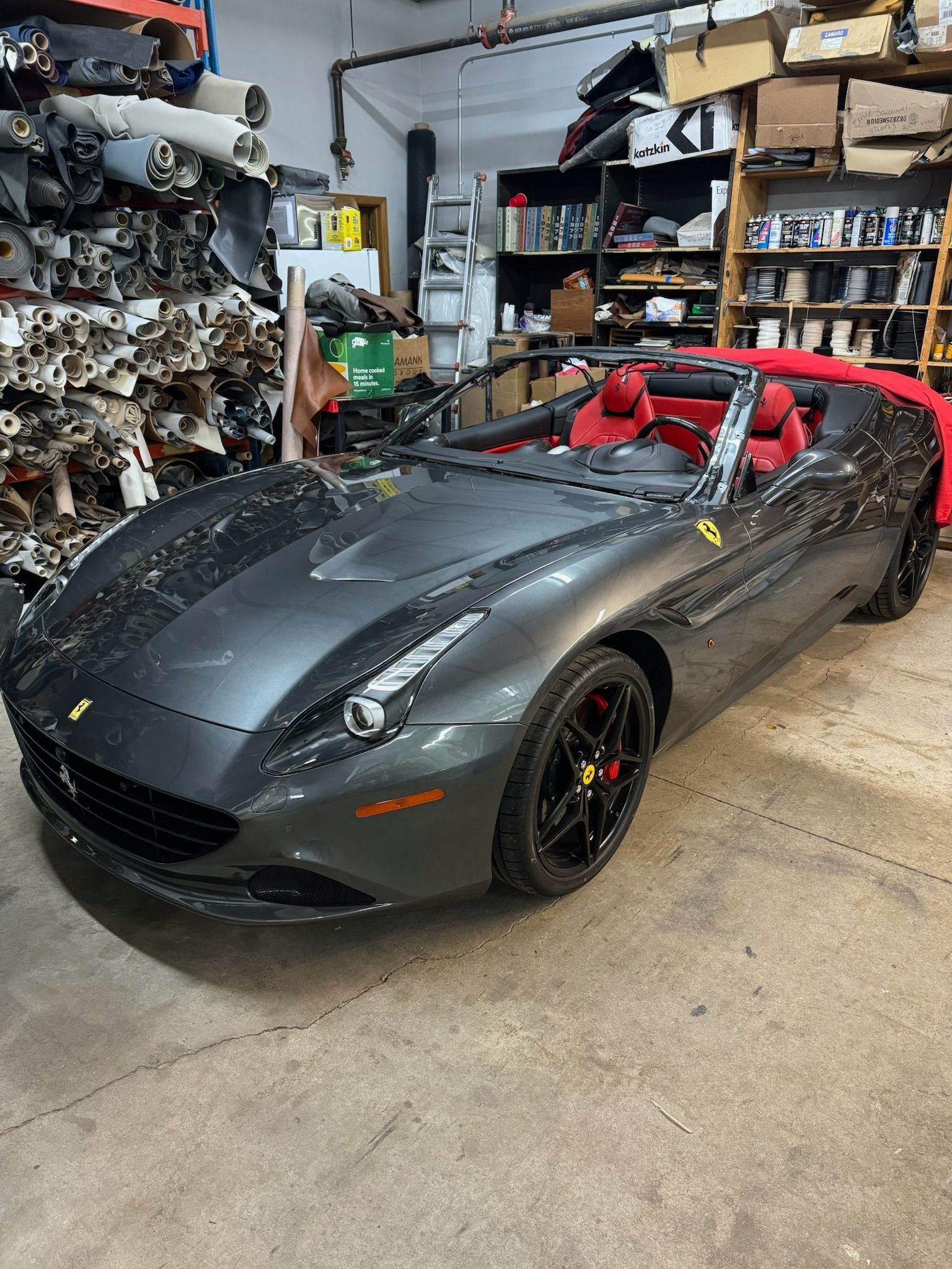 A gray Ferrari convertible with red leather seats parked inside a garage filled with car parts and storage shelving.