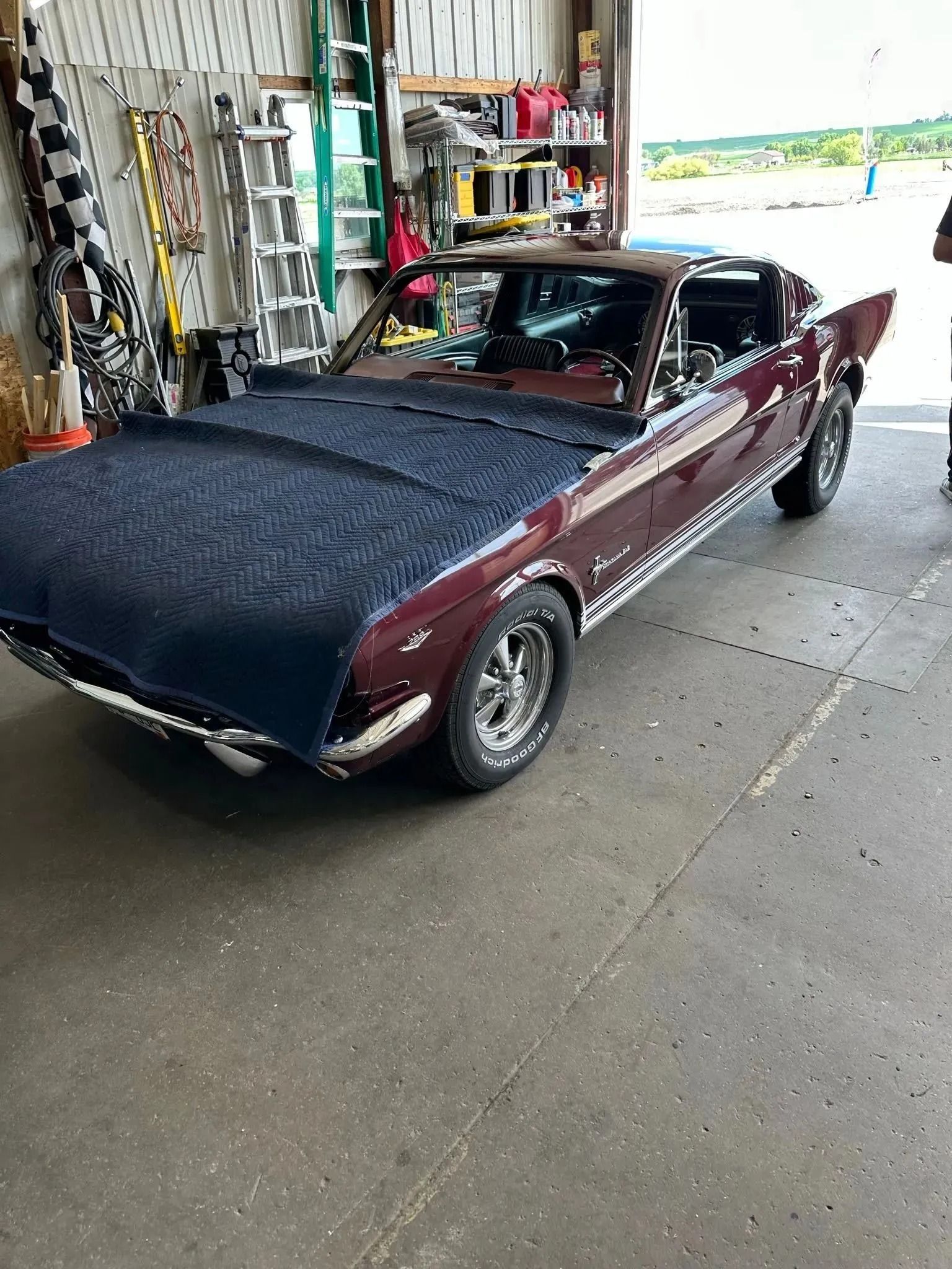 A dark maroon classic convertible parked inside a garage, with a blue quilted protective cover draped over its hood.