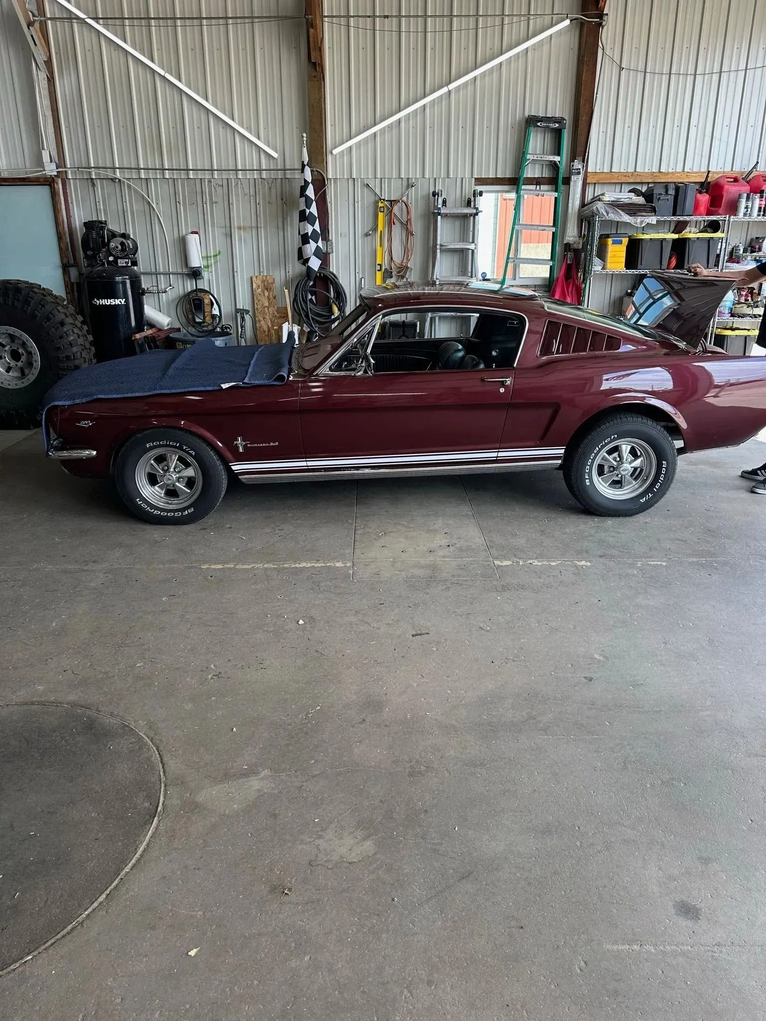 A deep red Ford Mustang fastback parked in a workshop with a blue cover on the hood and custom white side graphics.