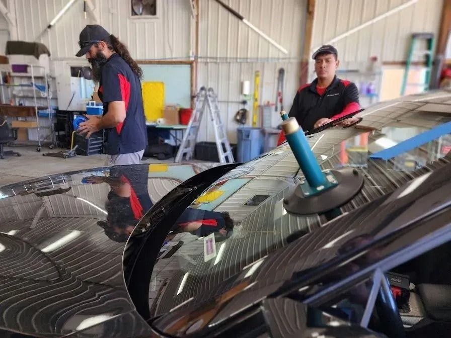 Two technicians in a garage work on replacing a car's windshield, which has a blue suction cup tool attached to it.