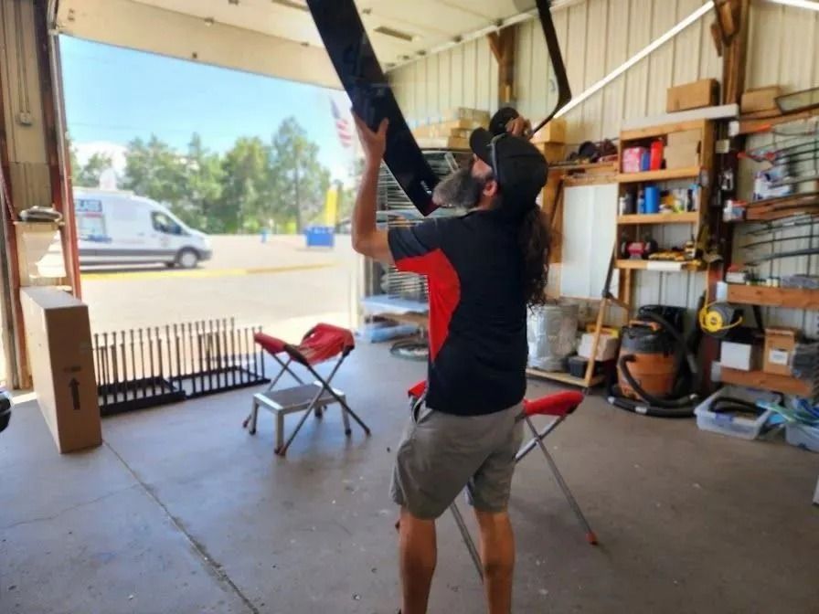 A person with long hair stands in a garage, lifting a long, dark component toward an open door.