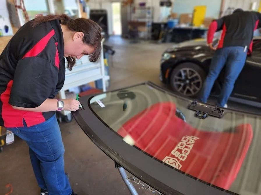 A person in a black and red uniform works on a windshield in an auto repair shop while another person works on a car.